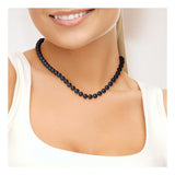 Une femme sourit, vêtue d'un haut blanc sans manches et portant le Perlinea Collier NOELYSE - un élégant collier en or blanc avec des perles noires de Tahiti de 7/8 mm - montré à partir des épaules sur un fond clair.