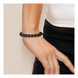 Une personne porte le bracelet Perlinea NOELYSE - un bracelet en perles de culture argentées et noires (9/10 mm) avec un fermoir boule - associé à un haut blanc et un pantalon noir, la main détendue le long du corps.