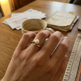 Bague or blanc 9 carats et perle de culture rose portée à l'index, posée sur une table en bois.