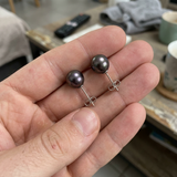 Puces d'oreilles en or blanc 9 carats avec perles de Tahiti grises, présentées dans une main.