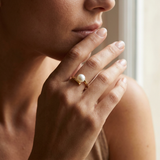 Bague or jaune 9 carats et perle de culture blanche portée à l'index, main levée près du visage.
