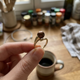 Bague or jaune 9 carats tenue entre les doigts, ornée d'une perle de culture chocolat.