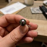 Boucle d'oreille bouton en or blanc 9 carats avec perle de Tahiti noire irisée, tenue entre les doigts.