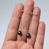 Boucles d'oreilles pendantes or blanc 9 carats et perles noires tenus dans une main.