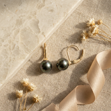 Dormeuses en or jaune 9 carats avec perles de Tahiti grises, posées sur lin beige.