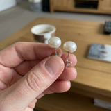 Puces d'oreilles en or blanc 9 carats serties de perles blanches, tenues entre les doigts.